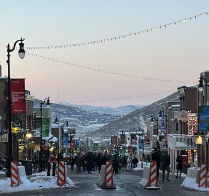 Main Street in Park City, Utah, is bustling with pedestrians during the 2025 Sundance Film Festival. Snow lines the sidewalks, festive string lights hang above the street, and colorful banners and storefronts glow under the early evening sky with mountains in the distance.