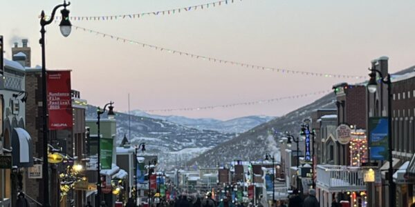 Main Street in Park City, Utah, is bustling with pedestrians during the 2025 Sundance Film Festival. Snow lines the sidewalks, festive string lights hang above the street, and colorful banners and storefronts glow under the early evening sky with mountains in the distance.