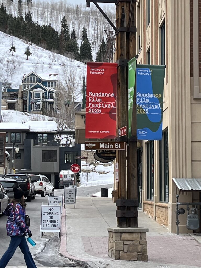 Banners for the Sundance Film Festival 2025 hang along Main Street in Park City, Utah, with snowy mountains and colorful houses in the background. Signs on the sidewalk warn against stopping or standing. A pedestrian in winter gear walks by, highlighting the festive yet wintry atmosphere.