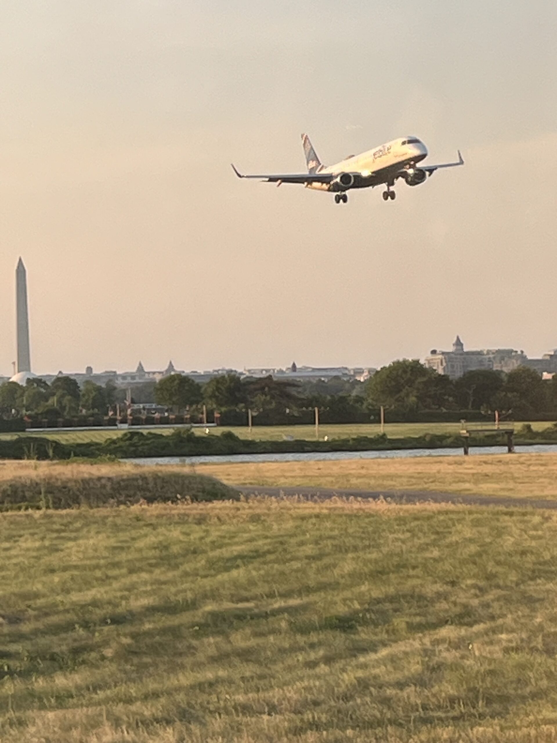 An airplane prepares to land at Reagan National Airport in Washington, D.C., during golden hour. The Washington Monument stands tall in the background, with soft evening light casting a warm glow over the grassy airfield and distant city skyline.