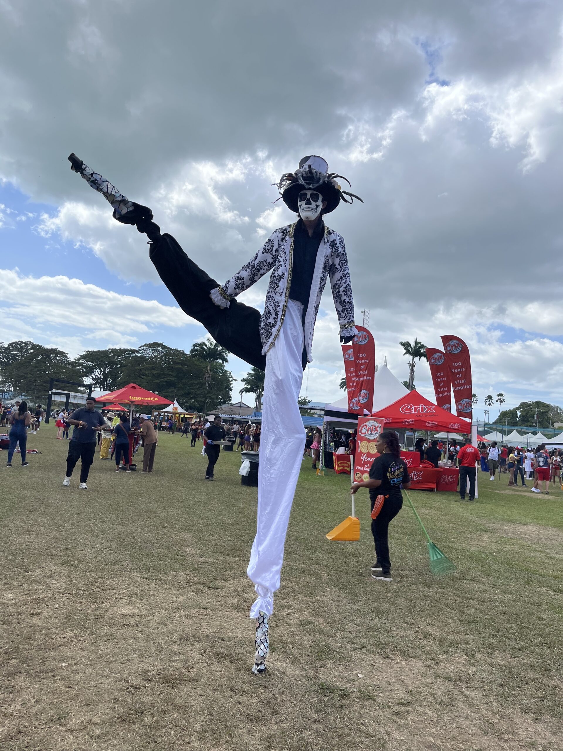 A costumed stilt walker performs at an outdoor festival in Trinidad, wearing dramatic white and black attire with a feathered top hat and skeleton face paint. One leg is lifted in a high kick. Carnival booths and people enjoying the event are visible in the background under a partly cloudy sky.