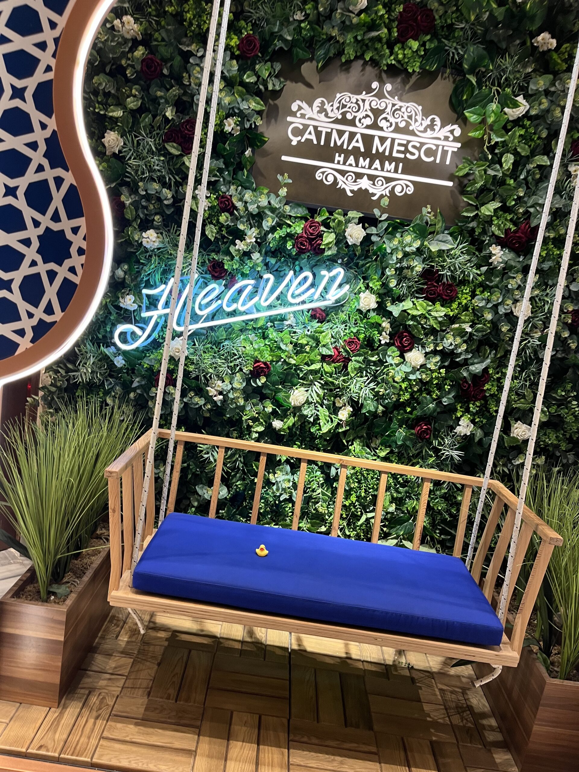 Wooden swing with a royal blue cushion and a small yellow rubber duck, set against a lush flower wall with a neon “Heaven” sign and the “Çatma Mescit Hamamı” logo, evoking a luxurious and whimsical spa entrance.