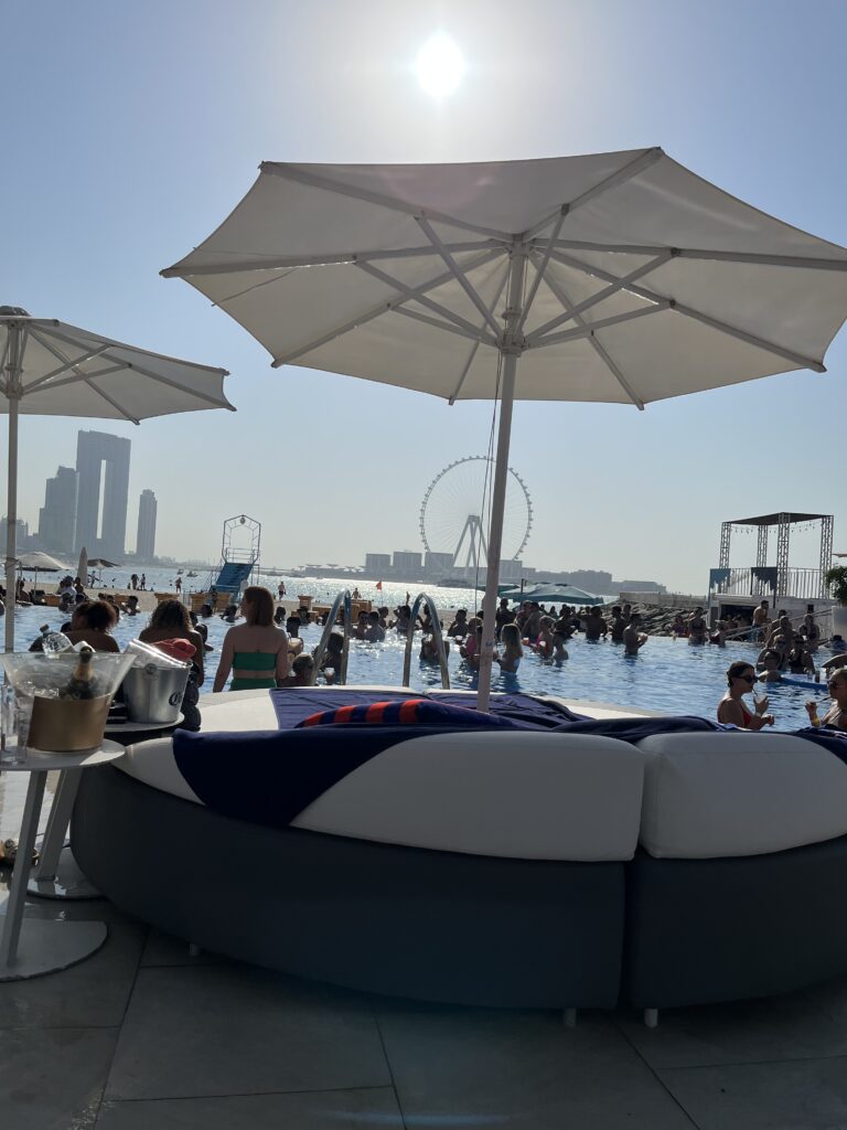 A sunny day at Zero Gravity beach club in Dubai featuring a crowded infinity pool with guests enjoying drinks and music. In the foreground, a large round poolside lounger is shaded by white umbrellas. The iconic Ain Dubai observation wheel and city skyline are visible in the background across the water.