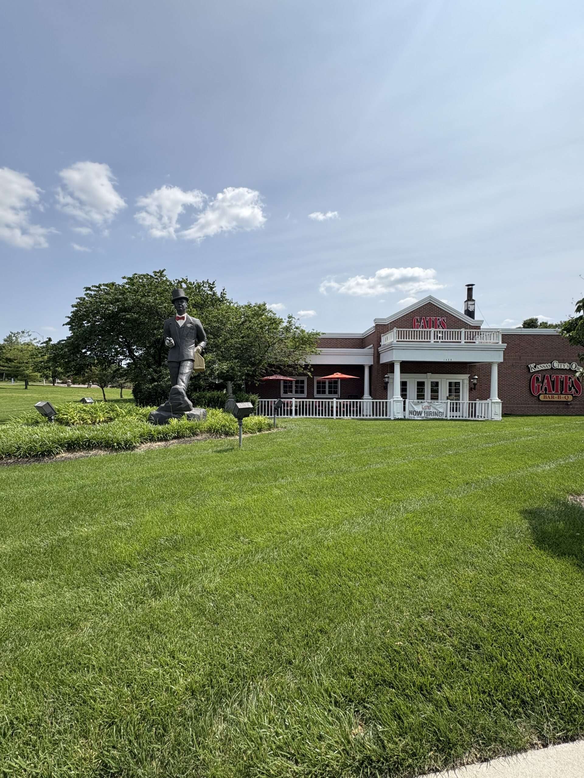 A bright, sunny day at Gates Bar-B-Q in Kansas City, Missouri. The image shows a large green lawn leading to the red-brick restaurant building with white trim and a sign reading "GATES" above the entrance. In front of the restaurant is a life-size statue of a man in a suit and bowler hat holding a briefcase and walking cane, a tribute to the restaurant’s founder.