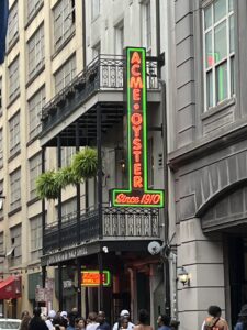 Vertical neon sign reading “ACME OYSTER Since 1910” glows brightly in red and green on the side of a historic French Quarter building in New Orleans. A small crowd waits outside beneath wrought iron balconies and hanging ferns, capturing the lively, classic atmosphere of Acme Oyster House.