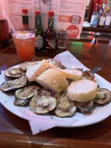 A sizzling plate of chargrilled oysters topped with melted cheese and garlic butter, served with slices of French bread and a cocktail at Acme Oyster House in New Orleans, with hot sauce bottles and a menu in the background.