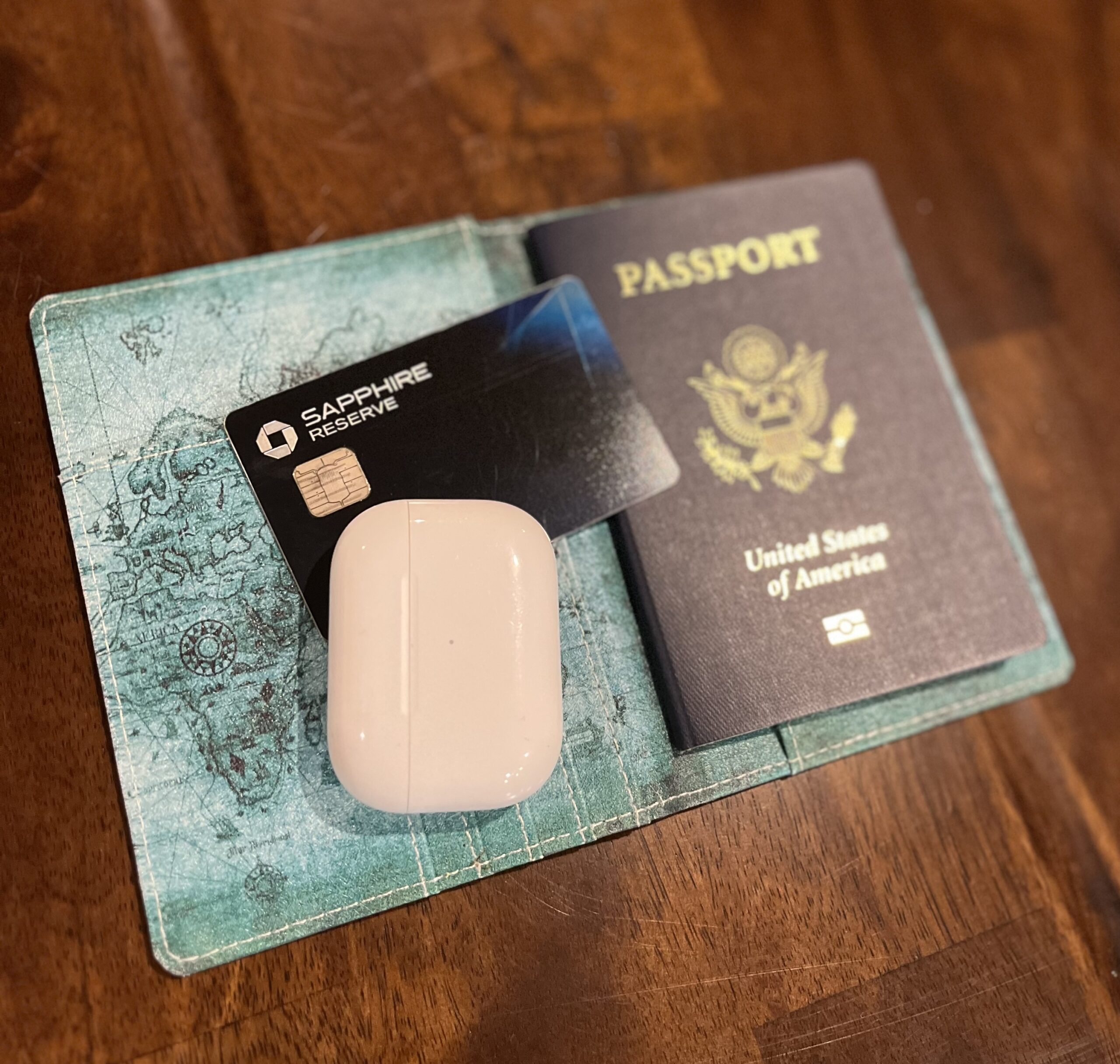 A travel essentials flat lay featuring a U.S. passport, a Chase Sapphire Reserve credit card, and a white AirPods case. All items are neatly placed on an aqua blue travel wallet with a vintage world map design, resting on a wooden surface.