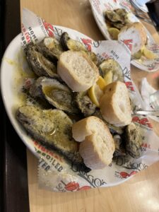 A plate of buttery, cheesy chargrilled oysters at Drago’s Seafood Restaurant, served with lemon wedges and French bread rounds. The oysters sit in pools of garlic butter on branded paper-lined plates.