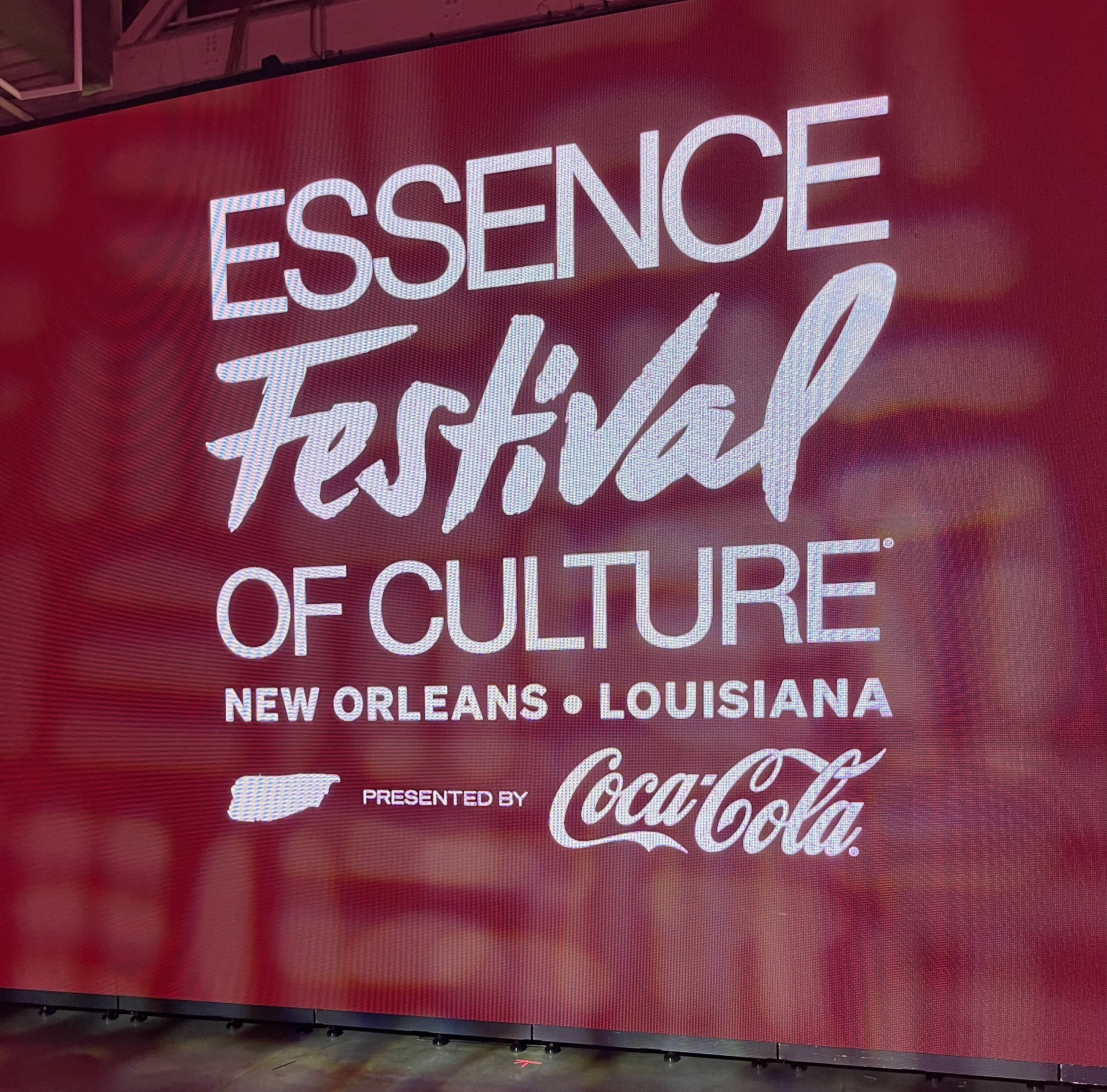 Large red digital screen at the Essence Festival of Culture 2025 in New Orleans, Louisiana, presented by Coca-Cola, featuring bold white text and event branding.