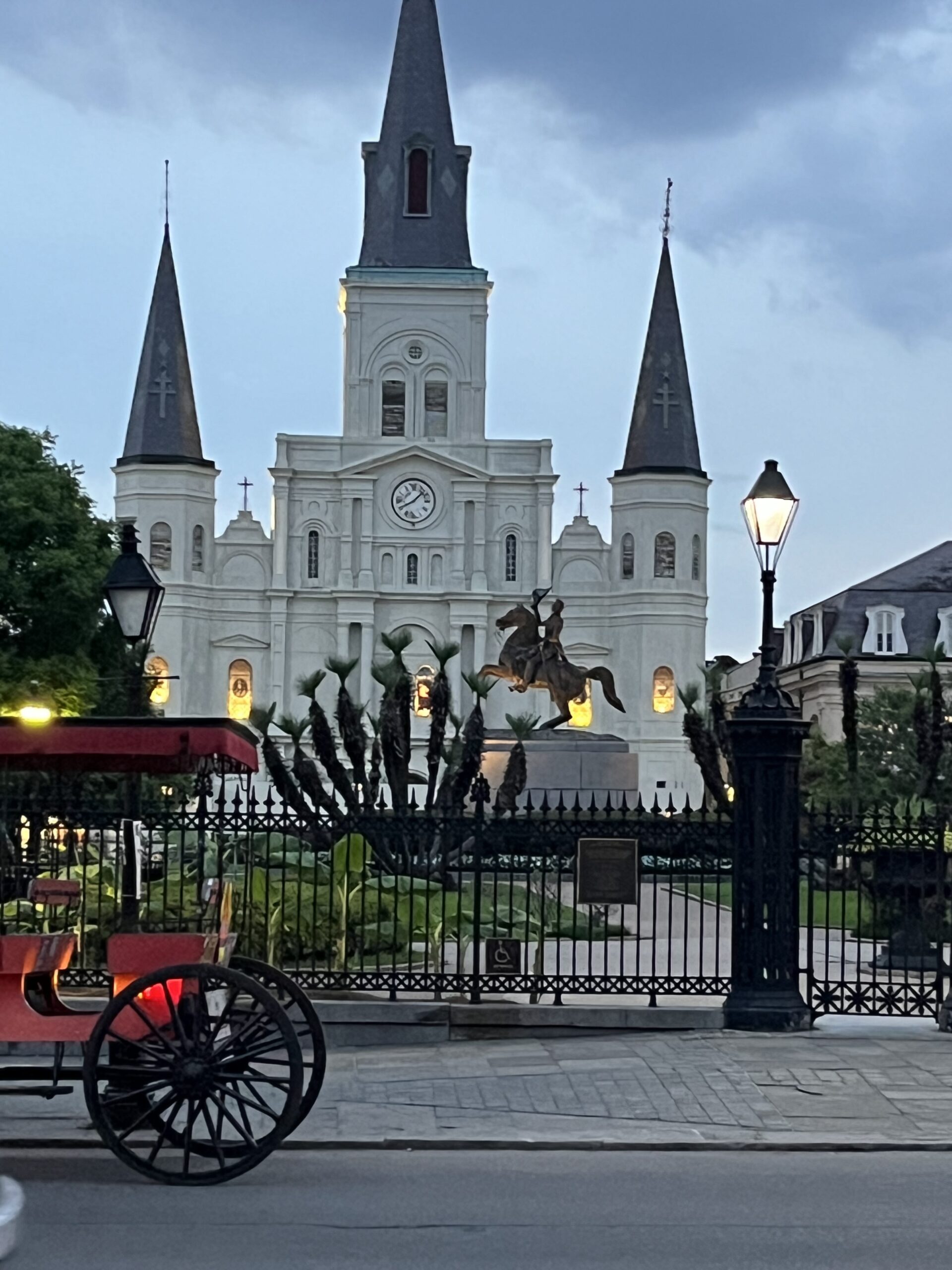 The iconic St. Louis Cathedral in New Orleans’ Jackson Square at dusk, flanked by gas lanterns and iron fences, with a red horse-drawn carriage in the foreground. A classic French Quarter scene.